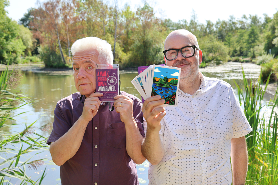 Two people outdoors near a pond, both holding colourful art postcards
